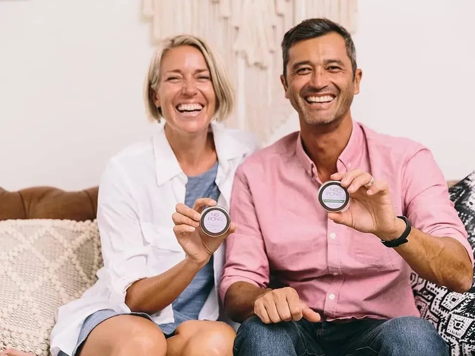 Melanie and Chris, the founders of No Pong, laughing on a couch and holding up their tins