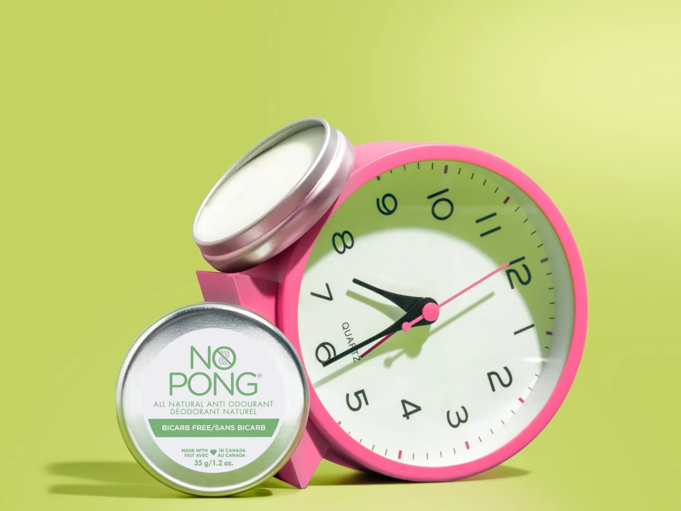 A No Pong Bicarb Free tin in front of a pink alarm clock, with an open tin on top, on a lime green background